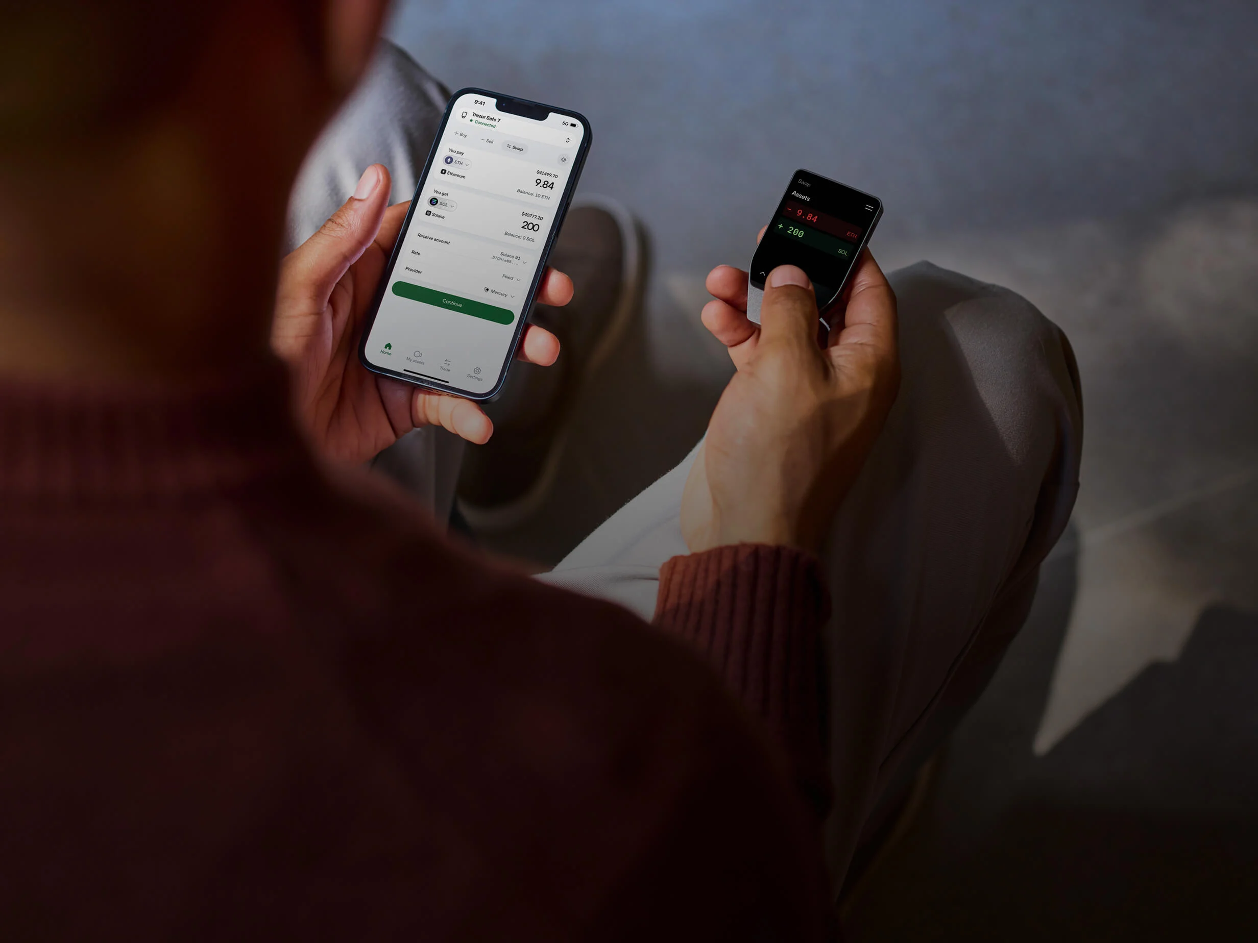 Blurry rear view of a man holding a smartphone in his left hand and a Trezor Safe 7 hardware wallet in his right hand.